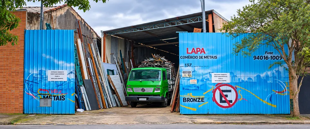 Fachada da Lapa Comércio de Metais com portões azuis pintados com o nome da empresa e o caminhão verde no pátio